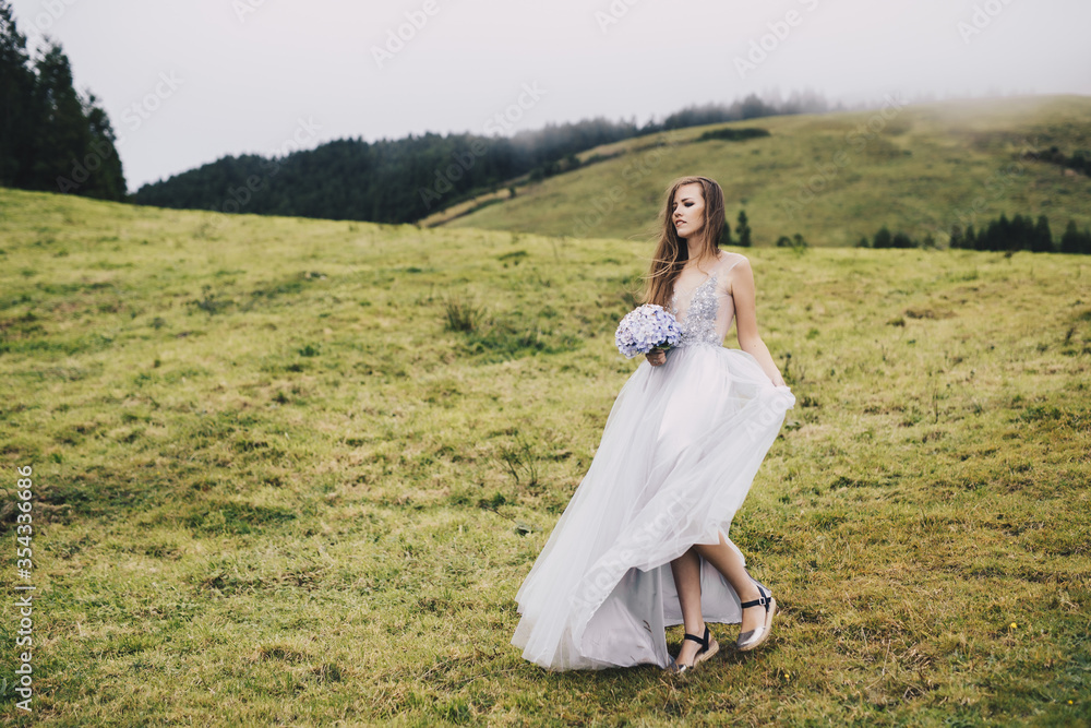 a young beautiful girl in a white dress looking like a bride walking through green meadows and hills with gartensia in her hands a young beautiful girl in a white dress looking like a bride walking through green meadows and hills with gartensia in her hands