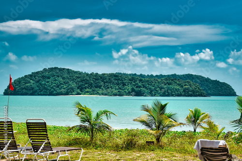 Small palm trees and deck chairs looking at a tropical island in the azure sea under the blue sky on the shores of the sandy beautiful exotic and stunning Cenang beach in Langkawi island