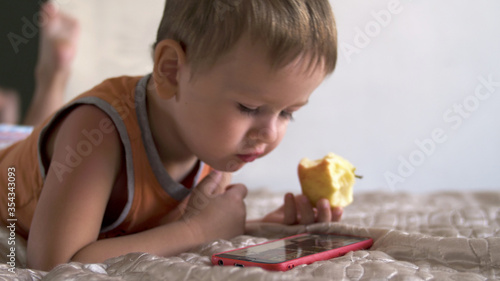 child boy eat apple and watch smartphone on bed at home. distance learning education