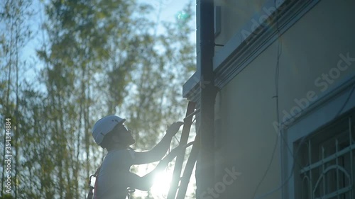 Young apprentice man at work, construction builder climbs up the ladder with safety equipment, white hardhat in construction site
