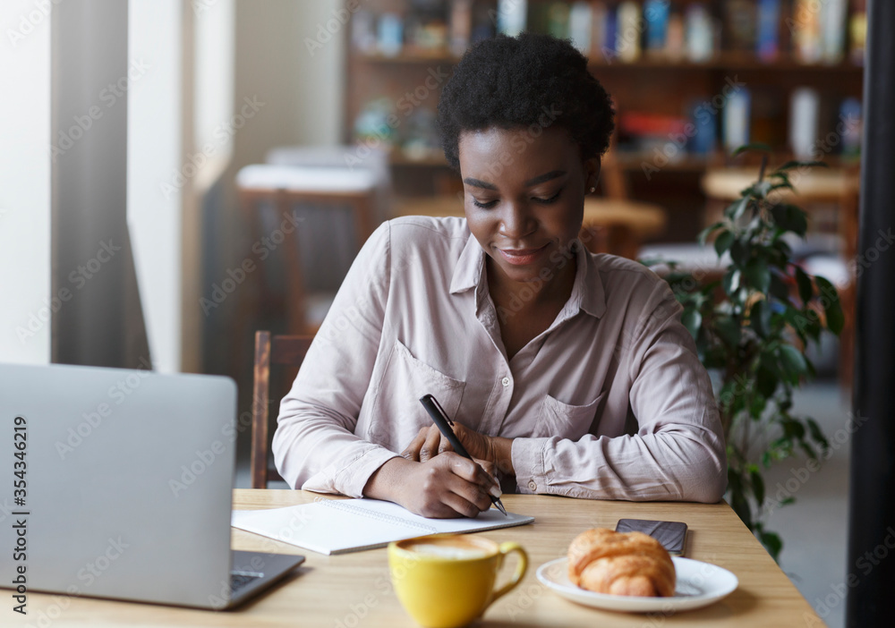 Studying online. Young African American girl with laptop taking notes while having breakfast in cafe