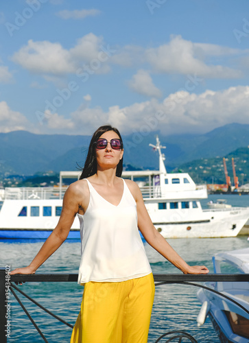 Positive woman walking at the harbor on vacation. Mountains on background. Cheerful, smiling, lucky woman in white blouse, yellow trousers and sunglasses, outdoors at sea port. Sunny summer day.