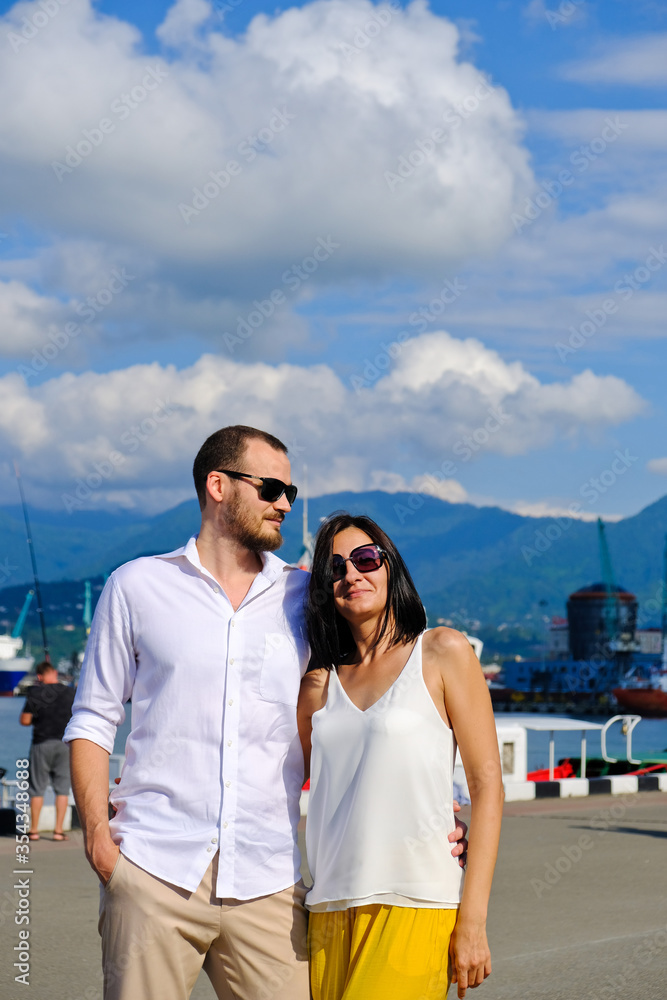 Happy couple of lovers walking at the harbour. Mountains at background. Cheerful, smiling man in white and woman in white blouse and yellow trousers, outdoors at sea port.