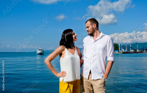 Happy couple of lovers walking at the harbour. Mountains at background. Cheerful, smiling man in white and woman in white blouse and yellow trousers, outdoors at sea port.