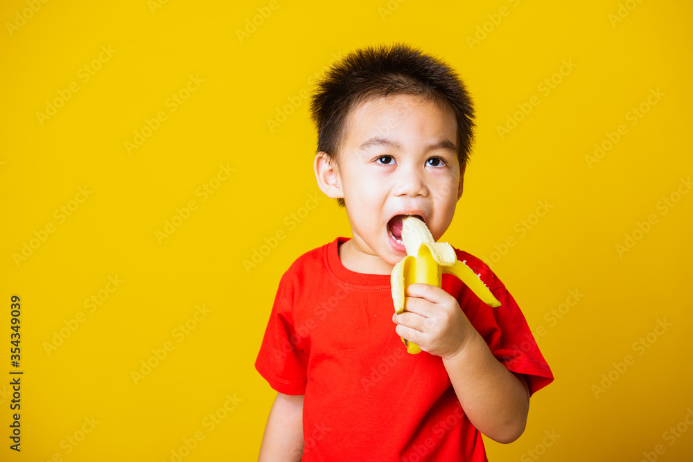 Happy portrait Asian child or kid cute little boy attractive smile wearing red t-shirt playing holds peeled banana for eating, studio shot isolated on yellow background Happy portrait Asian child or kid cute little boy attractive smile wearing red t-shirt playing holds peeled banana for eating, studio shot isolated on yellow background
