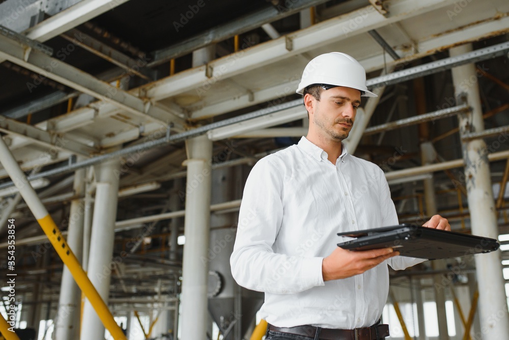 Close up of young Caucasian smiling worker with helmet on head using tablet for work while standing in warehouse. Close up of young Caucasian smiling worker with helmet on head using tablet for work while standing in warehouse.