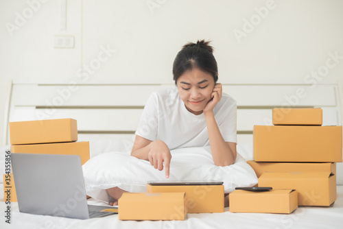 An Asian woman is using a tablet and a computer to happily find and choose products in her bed at home. Around her, there are a lot of boxes ordered from the online website.