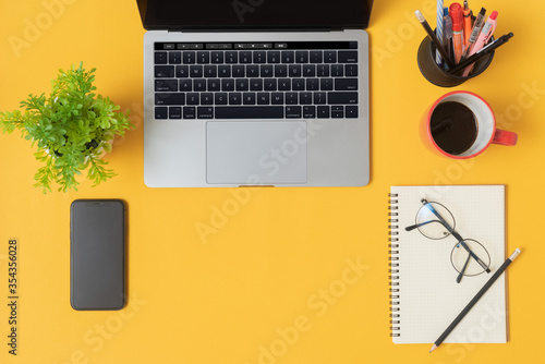 Orange and yellow desks with computers  coffee cups  pens  a notebook  pencils glasses   indoor ornamental plants  and a smartphone. This photo have copy space