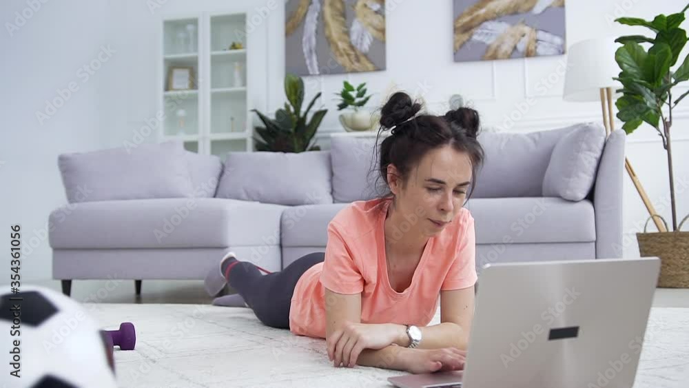 Attractive happy smiling young brunette in sportswear lying on the carpet in front of the laptop and doing stretching exercises using stretching strap Attractive happy smiling young brunette in sportswear lying on the carpet in front of the laptop and doing stretching exercises using stretching strap