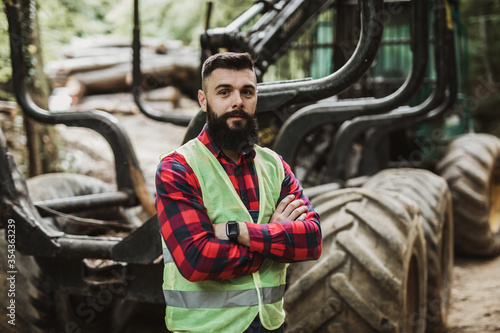 Young adult lumberjack or logger standing in front of huge bagger excavator in the woods and looking in the camera with crossed arms. photo