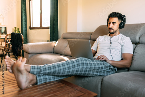 Man sitting in his pajamas on the couch at home working from a distance. The young man has the computer on his legs and the headphones on his head. I work from home