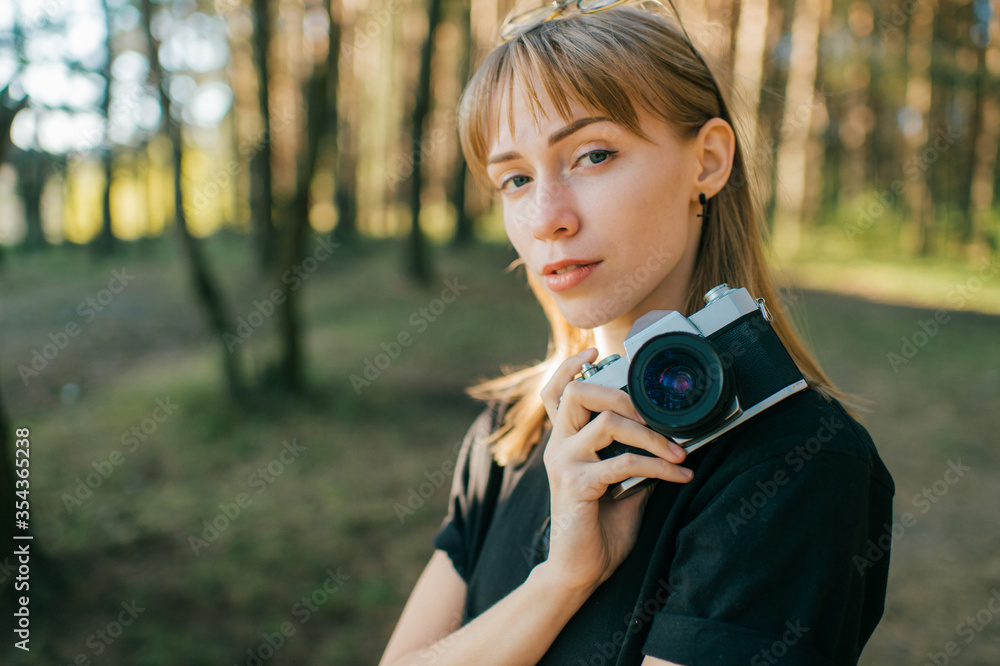 Portrait of beautiful young female photographer with short fair hair in black t-shirt holds a new camera and takes a lot of photos in the big forest in sunny weather Portrait of beautiful young female photographer with short fair hair in black t-shirt holds a new camera and takes a lot of photos in the big forest in sunny weather