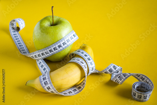 Green apple and banana with white tape measure on yellow background. Healthy lifestyle concept