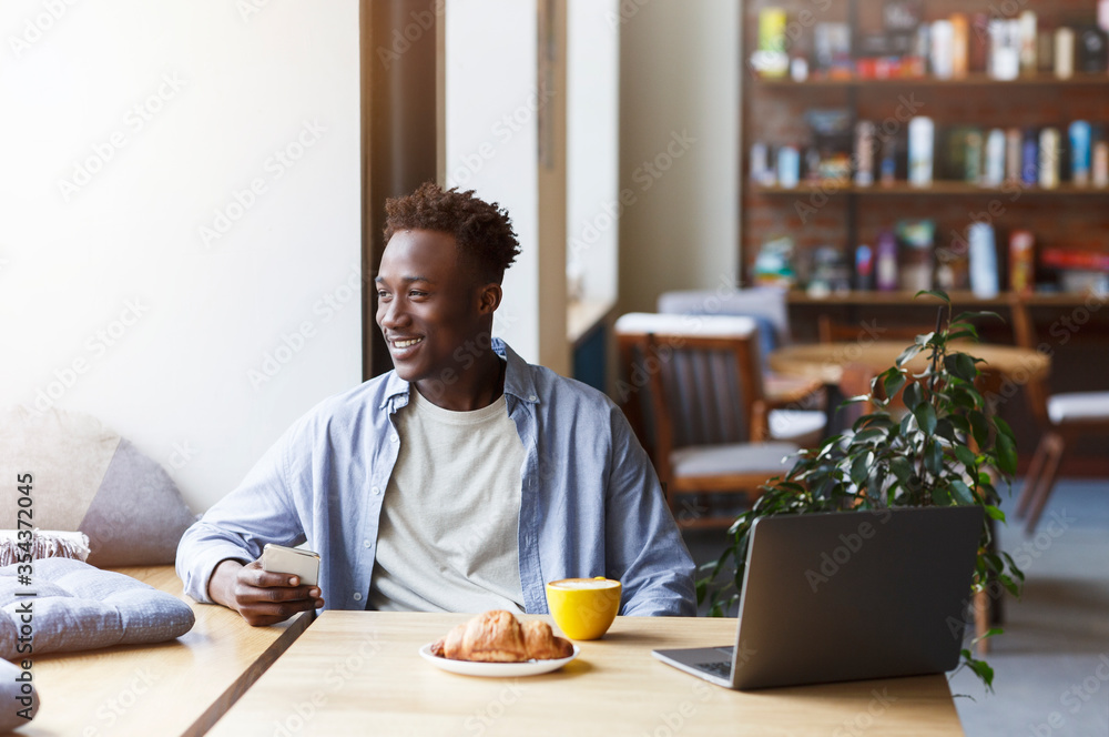 Millennial African American guy with smartphone and laptop enjoying morning coffee and croissant in cafe Millennial African American guy with smartphone and laptop enjoying morning coffee and croissant in cafe