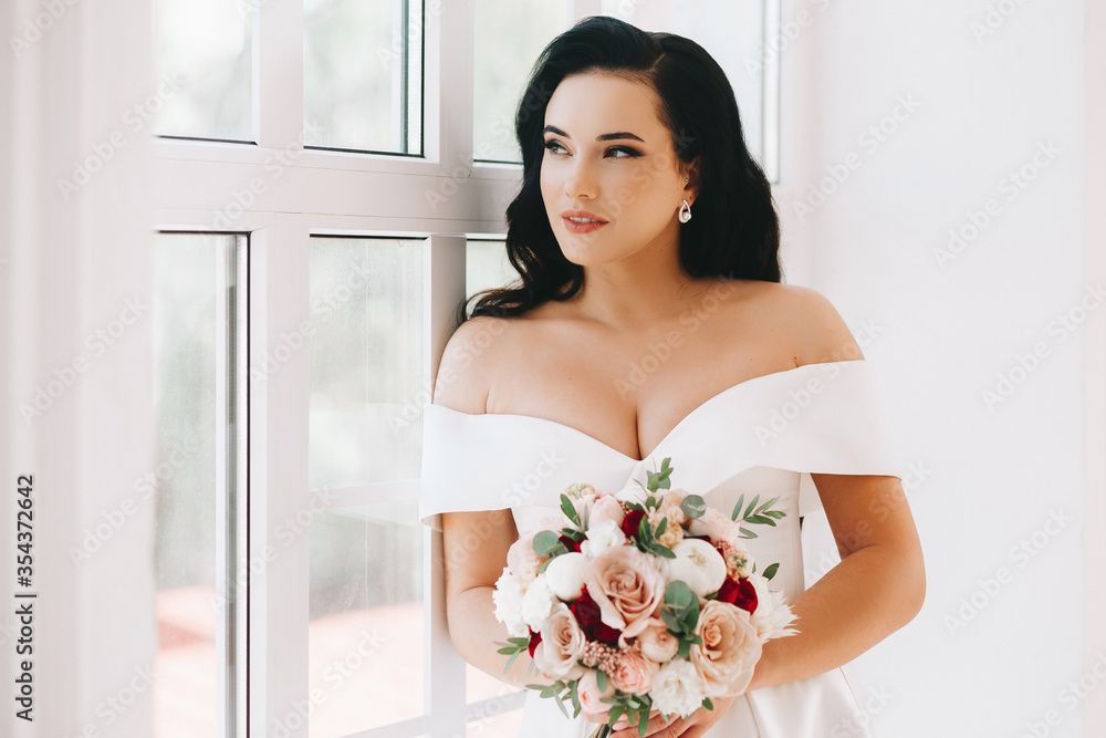 Beautiful bride is a young brunette of European appearance. Large single portrait with a bouquet of flowers. Wedding makeup and hairstyle. Model. In a bright studio by the window. Beautiful bride is a young brunette of European appearance. Large single portrait with a bouquet of flowers. Wedding makeup and hairstyle. Model. In a bright studio by the window.