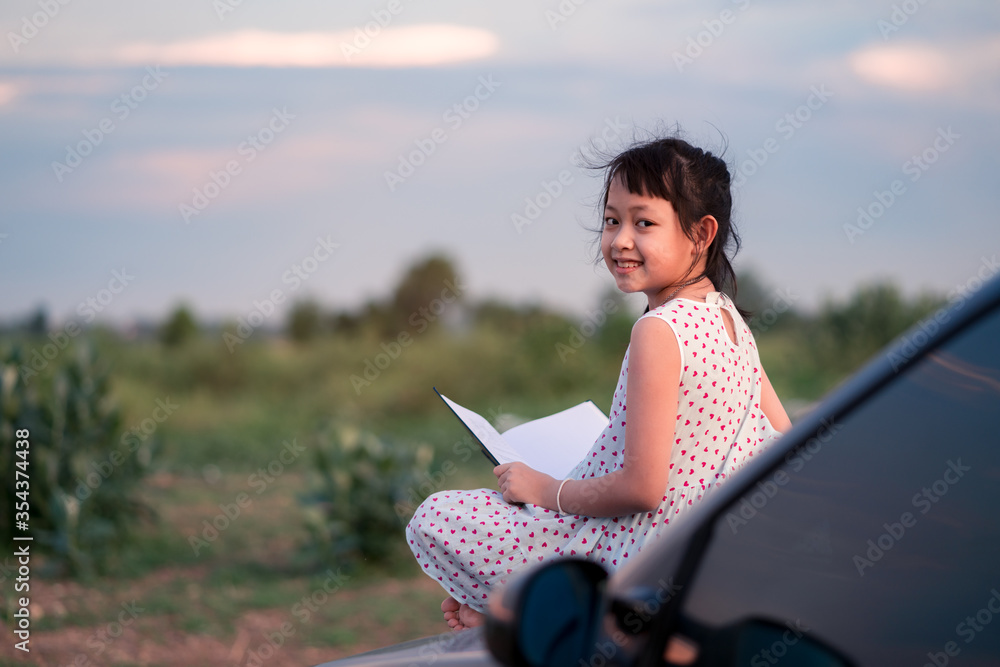 Little child girl  sitting and relaxing on a car reading a book in a green nature background