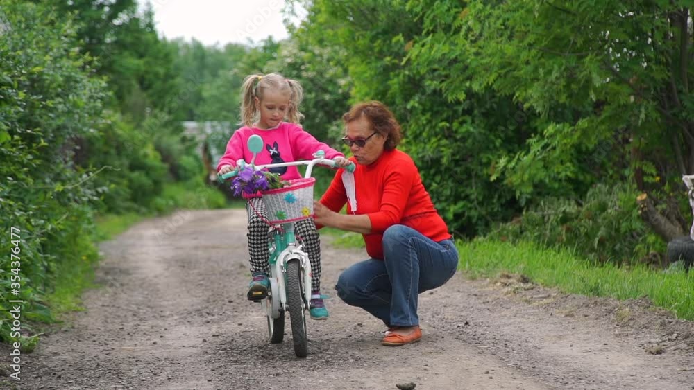 Caring Grandmother is Teaching Granddaughter How to Ride Bicycle. Little Girl Practicing Biking Outdoor. Slow Motion. Summer Sport Activities and Children Healthy Lifestyle Concept Caring Grandmother is Teaching Granddaughter How to Ride Bicycle. Little Girl Practicing Biking Outdoor. Slow Motion. Summer Sport Activities and Children Healthy Lifestyle Concept