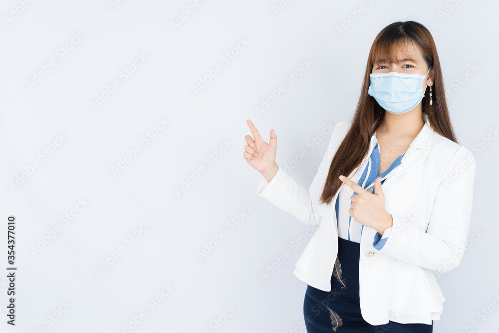Smiling Asian business woman wearing a medical face mask and pointing finger to side blank space over grey background. Back to the normal concept. Smiling Asian business woman wearing a medical face mask and pointing finger to side blank space over grey background. Back to the normal concept.