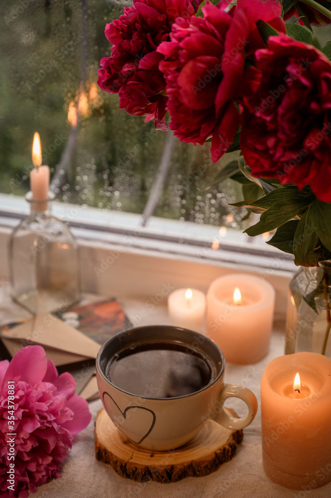 Cup with coffee on a windowsill with candles and flowers. Rain outside the window. Cup with coffee on a windowsill with candles and flowers. Rain outside the window.