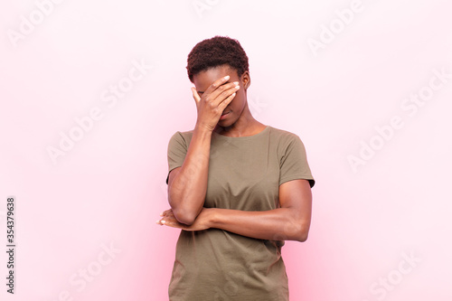 young pretty black womanlooking stressed, ashamed or upset, with a headache, covering face with hand against pink wall photo