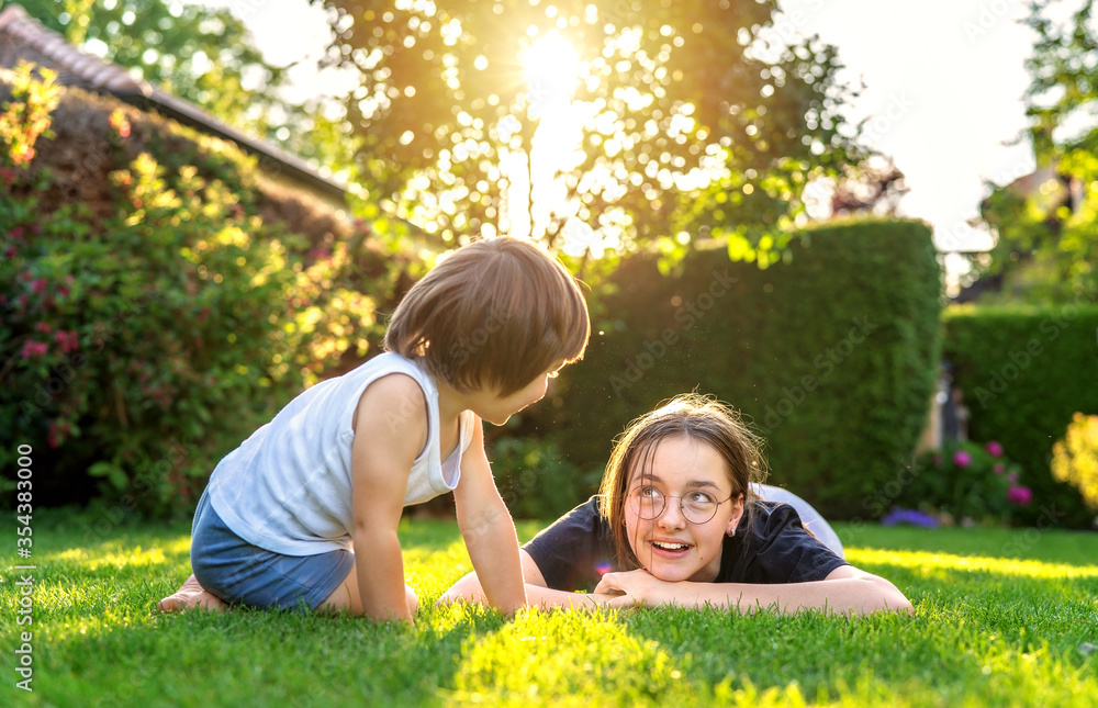 Fototapeta premium Happy siblings playing on green grass in garden backyard at sunset. Togetherness, little brother spending time with his teen sister. Summer weekend leisure outdoors activities. Childhood concept