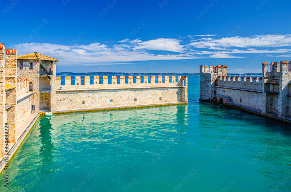 Small fortified harbor with turquoise water, Scaligero Castle Castello di Sirmione fortress, Sirmione town on Garda lake, medieval castle with stone towers and brick walls, Lombardy, Northern Italy