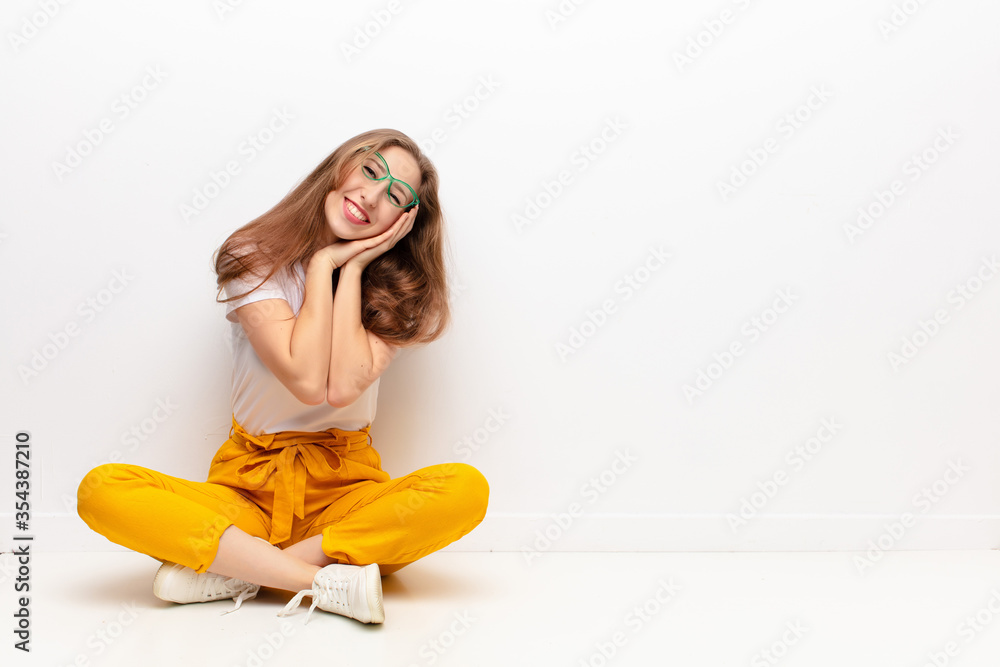 yound blonde woman feeling in love and looking cute, adorable and happy, smiling romantically with hands next to face sitting on the floor - obrazy, fototapety, plakaty 