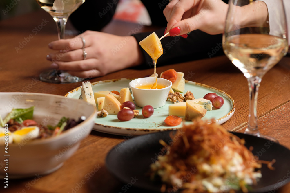 Female hand holding canape near cheese platter with different cheese, honey and grapes on a table for brunch. female friends - hands clinking white wine glasses, restaurant or bar on background, close