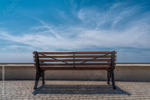 An empty bench against the background of blue sky and clouds in the Park stands on gray paving stones, in front of the parapet.