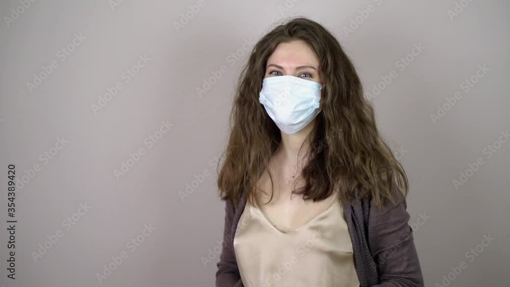 Brunette young woman putting on protective mask on face in coronavirus pandemic on grey background. Brunette young woman putting on protective mask on face in coronavirus pandemic on grey background.