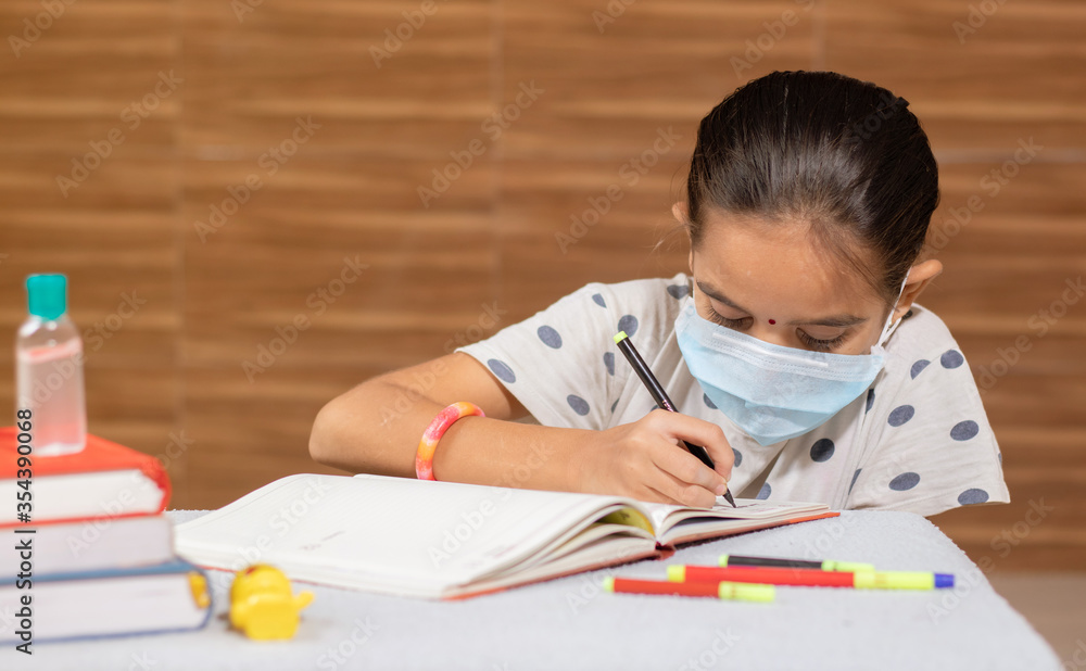 Concept of homeschooling, young girl with medical mask busy in writing at home during covid-19 or coronavirus pandemic lock down. Concept of homeschooling, young girl with medical mask busy in writing at home during covid-19 or coronavirus pandemic lock down.