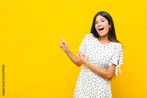 young pretty latin woman feeling joyful and surprised, smiling with a shocked expression and pointing to the side against flat wall