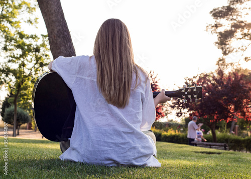 Canvastavla Rear view of young woman sitting on the grass and playing guitar in the park at