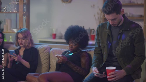  Slowmo tracking of young women and man sitting on couch and using mobile phones at boring house party