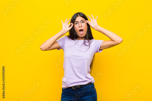 young pretty latin woman feeling shocked, amazed and surprised, holding glasses with astonished, disbelieving look against flat wall photo