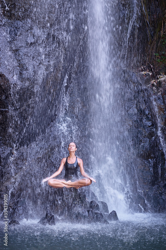 Wellness spa, vacation and yoga meditation concept. Pretty young woman in swimsuit sitting in lotus position on the rock under tropical waterfall.