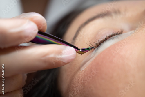 Woman lies on eyelash extension procedure in a beauty salon. Lashmaker holds tweezers with a bunch of artificial eyelashes. Close-up of a craftsman in gloves.
