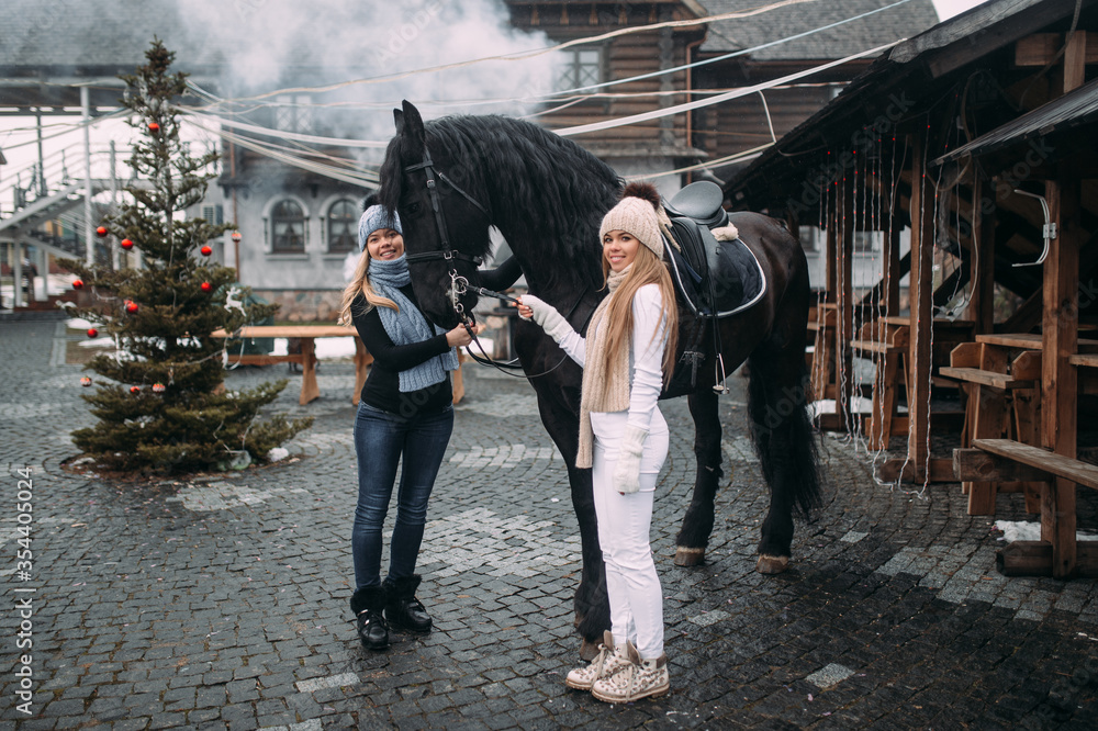 Beautiful caucasian girls with fair hair in warm clothes and knitted hats walks with a big black horse on the street