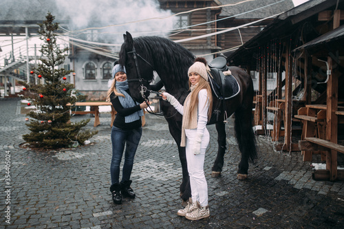 Beautiful caucasian girls with fair hair in warm clothes and knitted hats walks with a big black horse on the street
