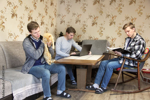 The same man. A man holds a cat in his arms. A man works at a laptop. A man in a chair reading a book. The cloned man. Three men of the same kind are busy. Doing many things at once