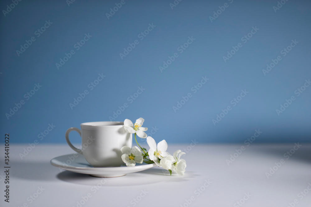 White cup of morning espresso with a blossoming apple tree branch on a stone surface of a table with reflections opposite blue background. White cup of morning espresso with a blossoming apple tree branch on a stone surface of a table with reflections opposite blue background.