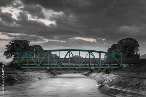 Black and white,  golden and green only, motion blur long exposure train passing over metallic railway bridge during blue hour with sky still dramatic after sunset photo