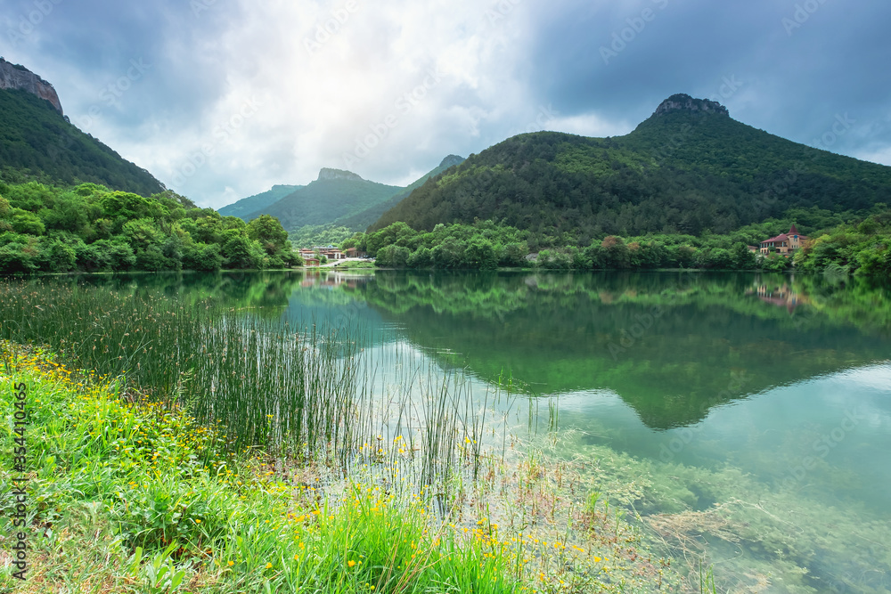 The lake is surrounded by greenery and mountains. Clear green water reflects the peaks. The sun breaks through the clouds.