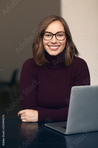 Portrait of beautiful smiling female student wearing glasses, sitting in front of her laptop, looking Confident