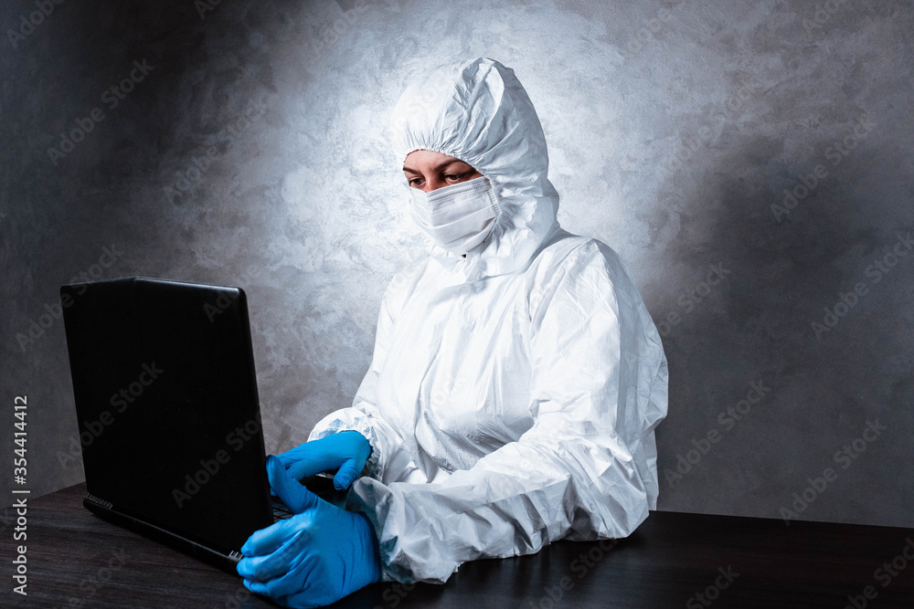 A virologist in a white mask and medical gloves dressed in a white protective suit works with a laptop and looks for information about coronavirus. Isolated and in panic due to epidemic. A virologist in a white mask and medical gloves dressed in a white protective suit works with a laptop and looks for information about coronavirus. Isolated and in panic due to epidemic.