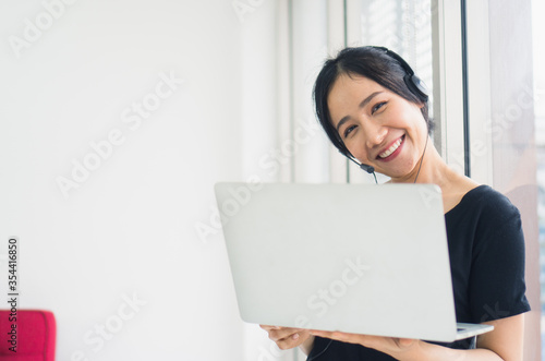 Young beautiful Asian woman use a headset working at home meeting by notebook standing look camera in living room smiling felling happy. corona virus, COVID-19 Pandemic new normal life at home Concept