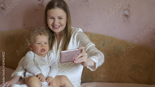Beautiful attractive mother with a cute baby sits at home on the couch and gazes cheerfully at a mobile phone.