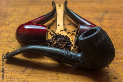 Wooden spoon with a heap of tobacco blend in the middle of three classic red, black and brown smoking pipes on a wooden yellow table.