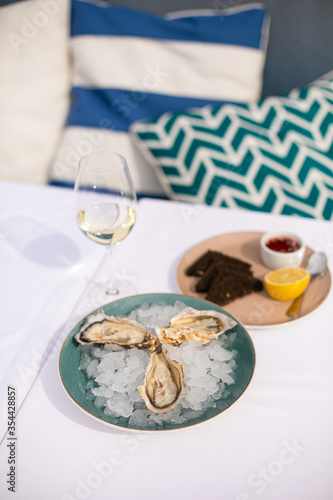 fresh seafood. Oysters lie on a plate with ice. In the background is a glass of white wine. A plate with sauce and vinegar and black croutons. White tablecloth. A restaurant. Blue pillows