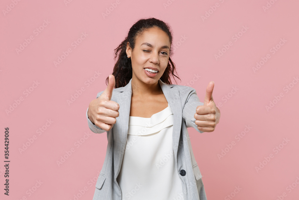 Blinking young african american business woman in grey suit, white shirt isolated on pink background studio portrait. Achievement career wealth business concept. Mock up copy space. Showing thumbs up. Blinking young african american business woman in grey suit, white shirt isolated on pink background studio portrait. Achievement career wealth business concept. Mock up copy space. Showing thumbs up.